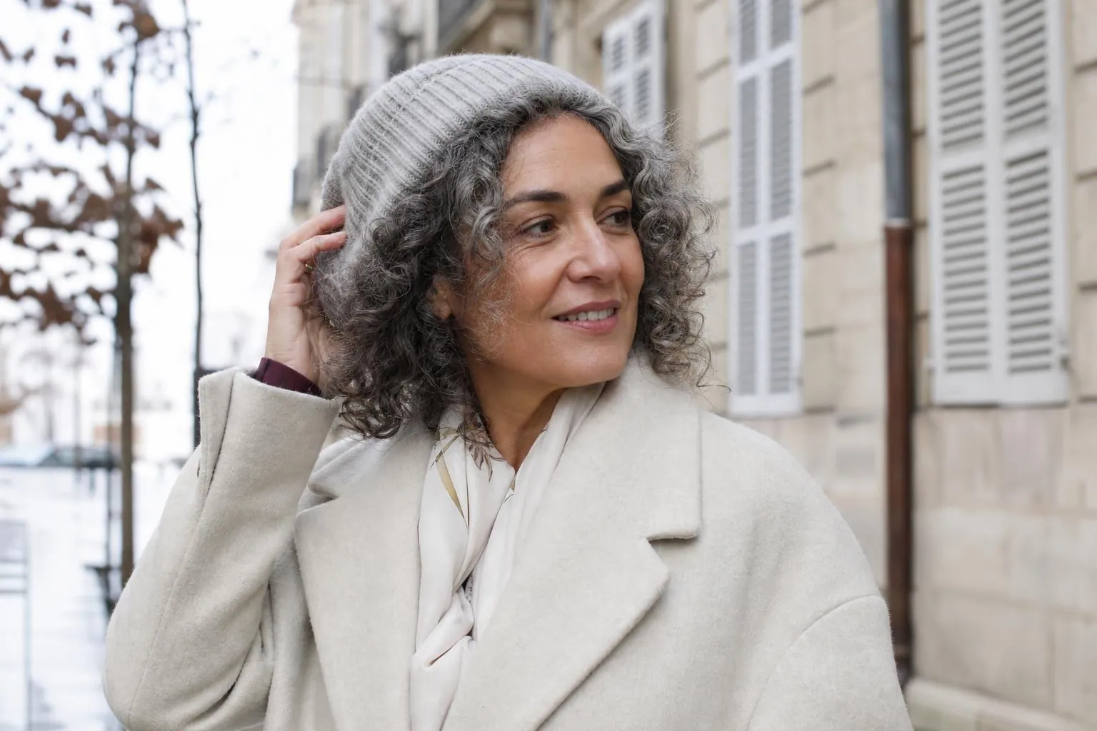 Femme d’une cinquantaine d’années en manteau de laine dans une rue française, boucles sorties d’un bonnet, lumière d’automne
