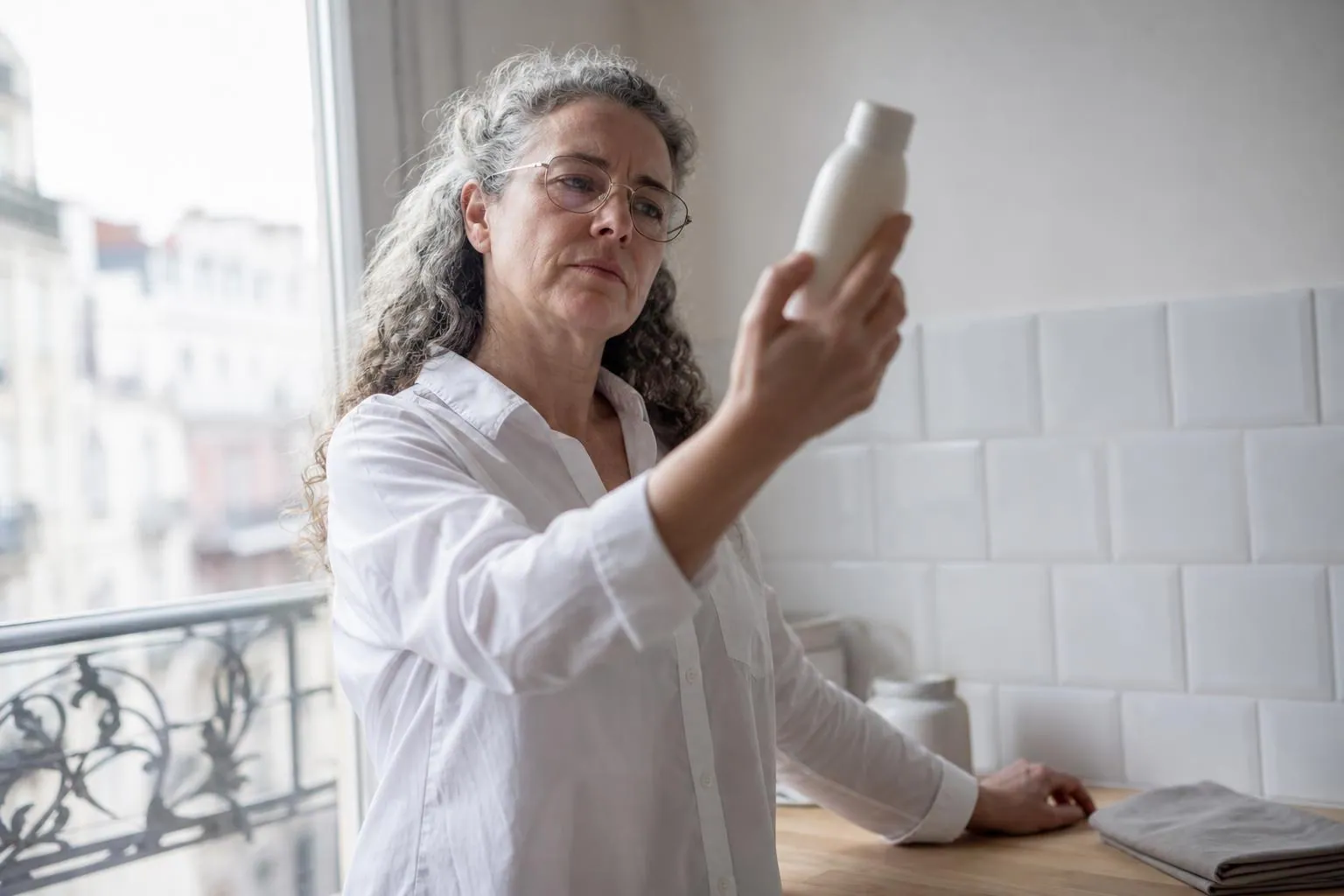 Femme d’une cinquantaine d’années lisant la composition au dos d’un flacon neutre près d’une fenêtre de cuisine