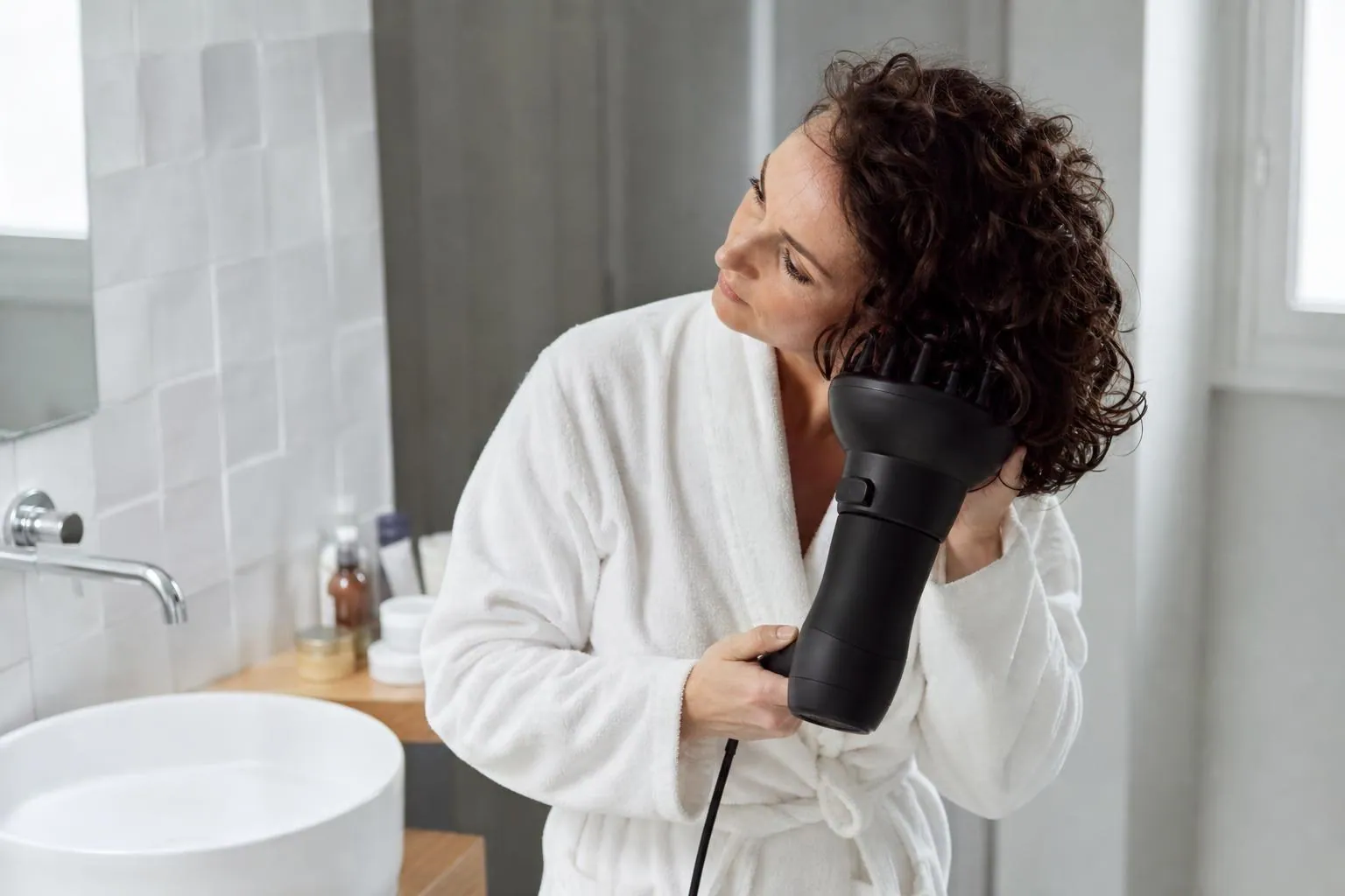 Femme d’une quarantaine d’années séchant ses cheveux bouclés au diffuseur, tête inclinée sur le côté, dans une salle de bain claire
