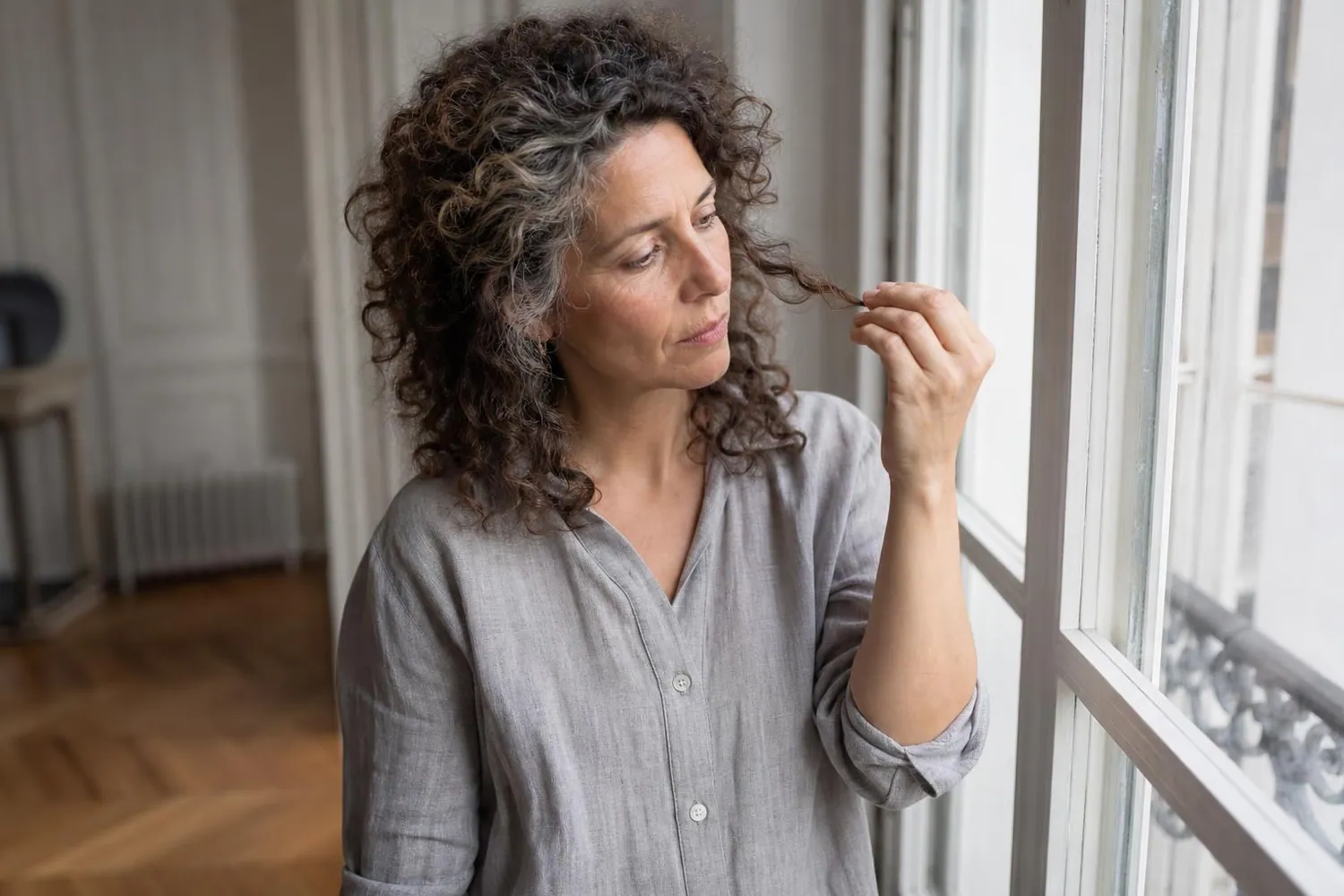 Femme d une cinquantaine d annees aux cheveux boucles poivre et sel soulevant une meche pres d une fenetre, lumiere naturelle